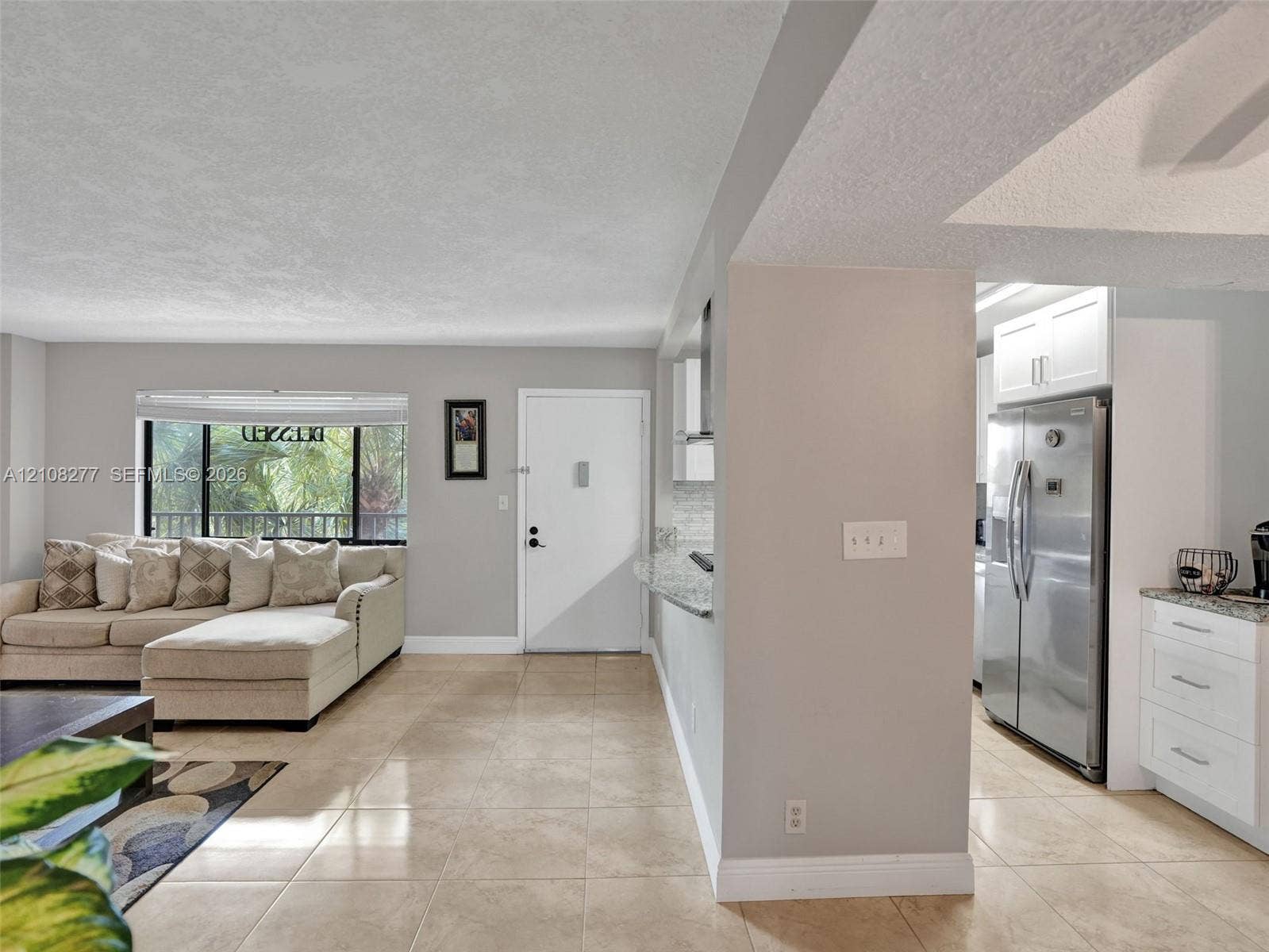 7466 Ashmont Circle, #303, Tamarac, FL 33321 Open-concept interior featuring large-format tile flooring, recessed kitchen cabinetry, stainless steel refrigerator, and oversized window with exterior greenery
