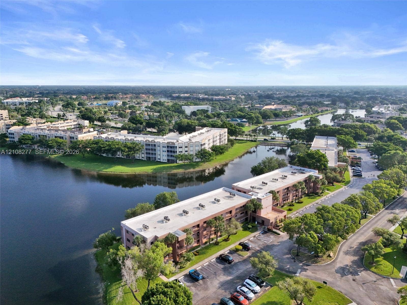 7466 Ashmont Circle, #303, Tamarac, FL 33321 Multi-story residential complex featuring waterfront positioning, expansive exterior parking, manicured landscaping, and mature trees