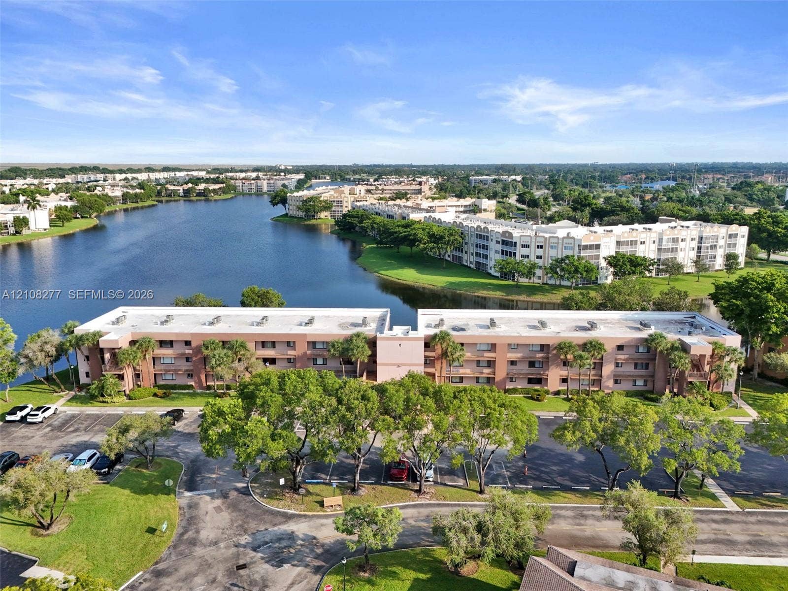 7466 Ashmont Circle, #303, Tamarac, FL 33321 Multi-story residential building featuring stucco exterior, expansive lake frontage, mature trees, and dedicated surface parking