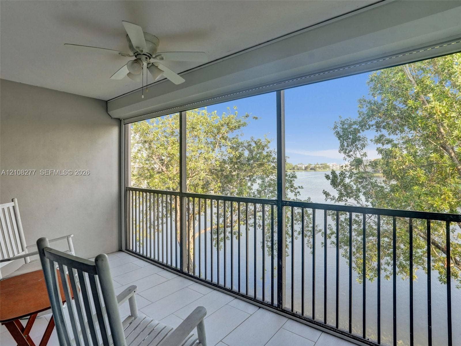 7466 Ashmont Circle, #303, Tamarac, FL 33321 Screened balcony featuring tile flooring, a ceiling fan, and metal railings overlooking a lake and surrounding trees