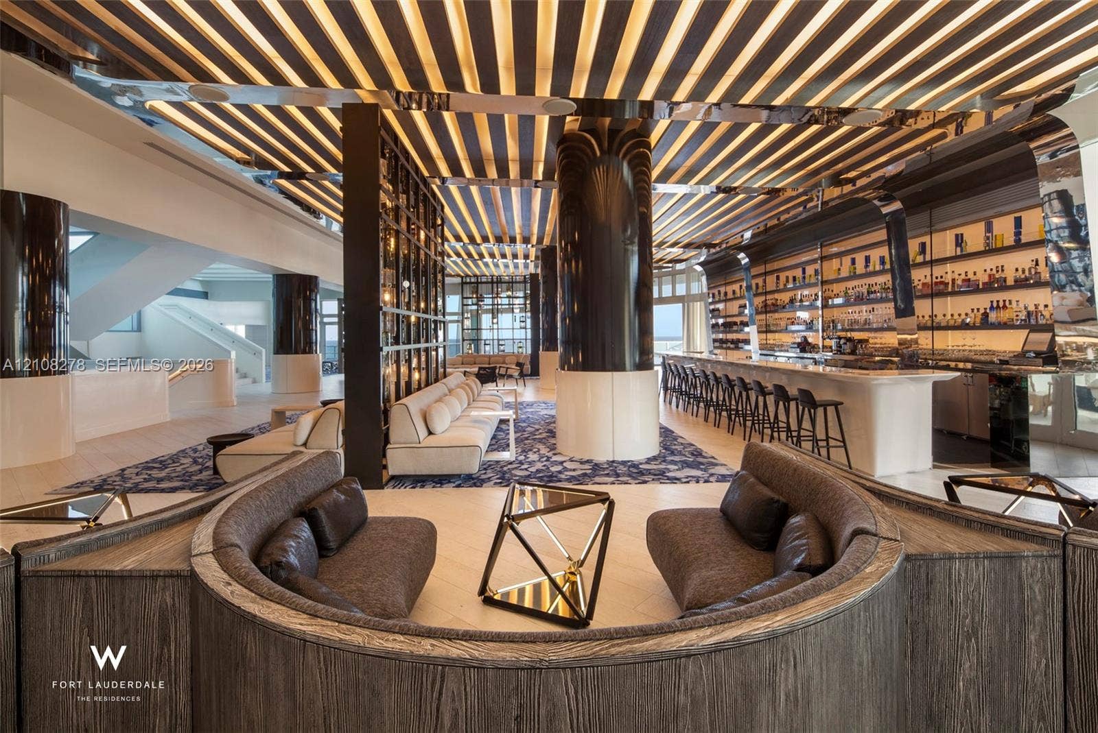 3101 Bayshore Drive, #701, Fort Lauderdale, FL 33304 Linear coffered ceiling featuring integrated lighting, custom bar with stone-finish surfacing, curved wood-paneled seating nook, polished architectural columns, and expansive shelving