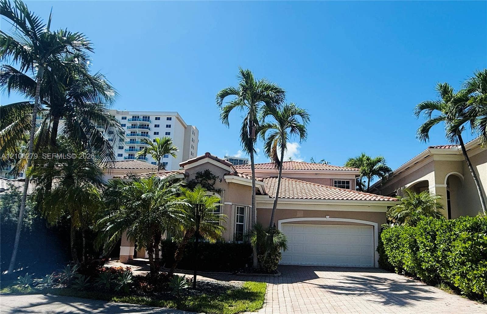 19424 39th Avenue, Sunny Isles Beach, Sunny Isles Beach, FL 33160 Stucco exterior residence featuring a multi-pitched clay tile roof, arched garage entrance, and paved driveway surrounded by mature palm trees and manicured hedging
