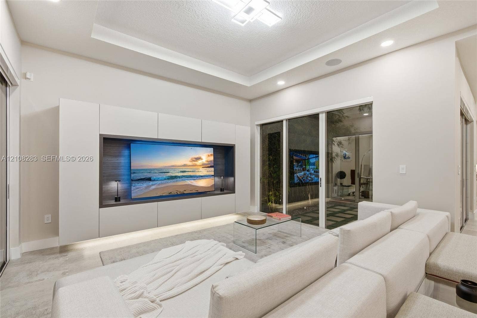 15215 SW 176th Lane, Miami, FL 33187 Living area featuring a tray ceiling with recessed lighting, custom floor-to-ceiling built-in media cabinetry with integrated lower illumination, and glass sliding doors