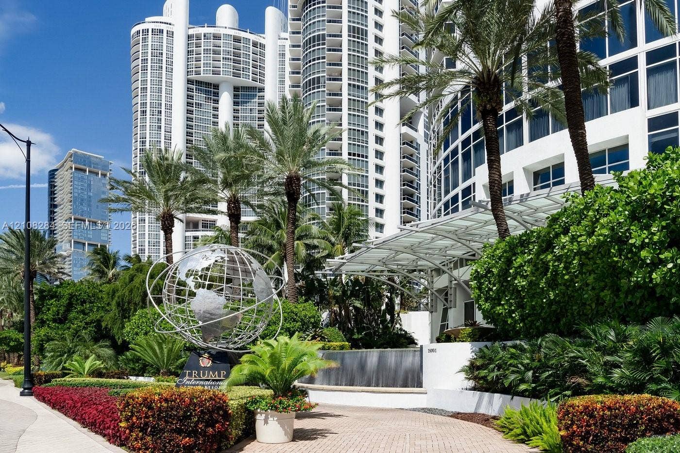 18001 Collins Avenue, #908, Sunny Isles Beach, FL 33160 High-rise building exterior featuring a white curved facade, metal-frame porte-cochre, decorative metal globe sculpture, tiered water feature, and mature tropical landscaping