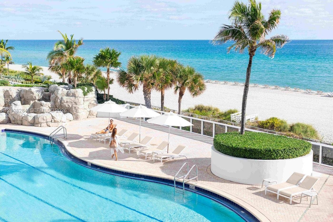 18001 Collins Avenue, #908, Sunny Isles Beach, FL 33160 Curved swimming pool featuring a stone-faced grotto, expansive paver pool deck, elevated circular planter with mature palm tree, and white perimeter fencing