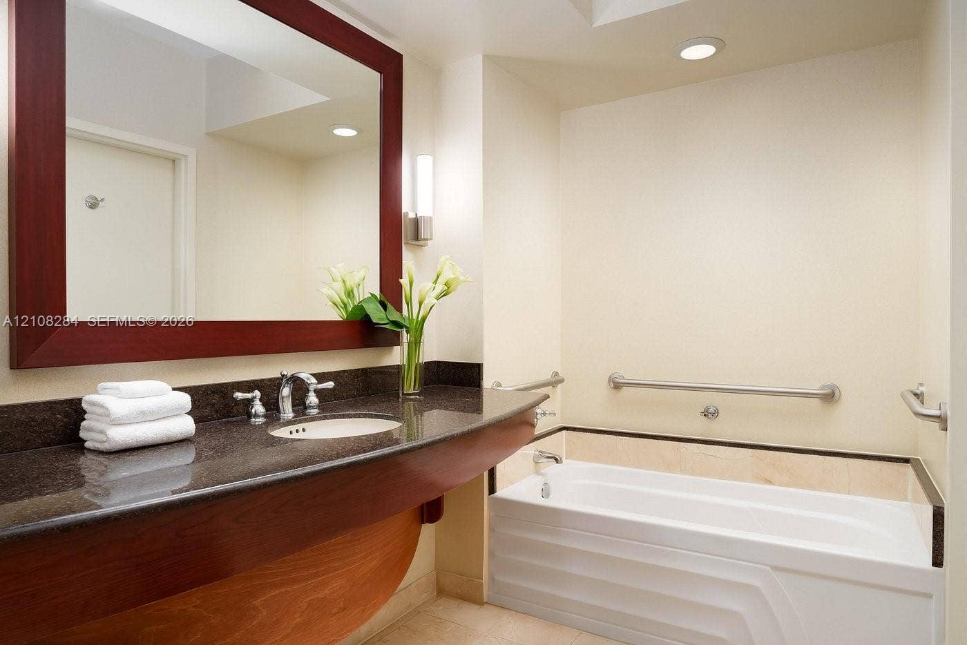 18001 Collins Avenue, #908, Sunny Isles Beach, FL 33160 Floating wood vanity with dark stone countertop, integrated sink, large framed mirror, and bathtub with wall-mounted grab bars