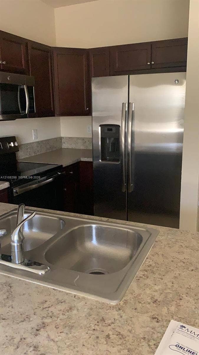 Kitchen featuring dark-finish cabinetry, stainless steel side-by-side refrigerator, integrated microwave, dual-basin stainless steel sink, and stone-finish countertops