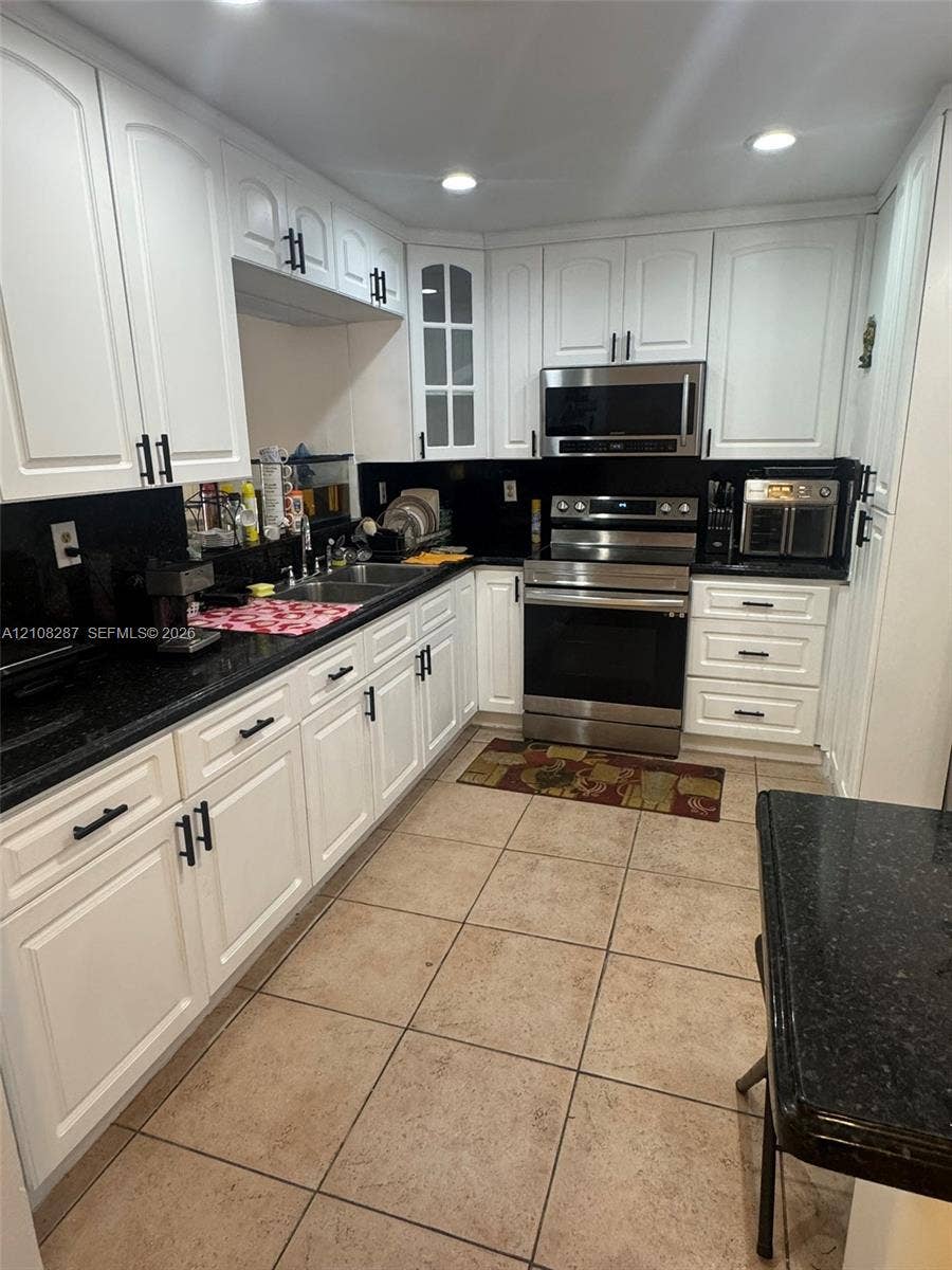 5350 NW 183rd Street, Miami Gardens, FL 33055 Kitchen featuring white shaker-style cabinetry with matte black hardware, dark stone countertops, stainless steel appliances, glass-panel upper cabinet doors, and tile flooring