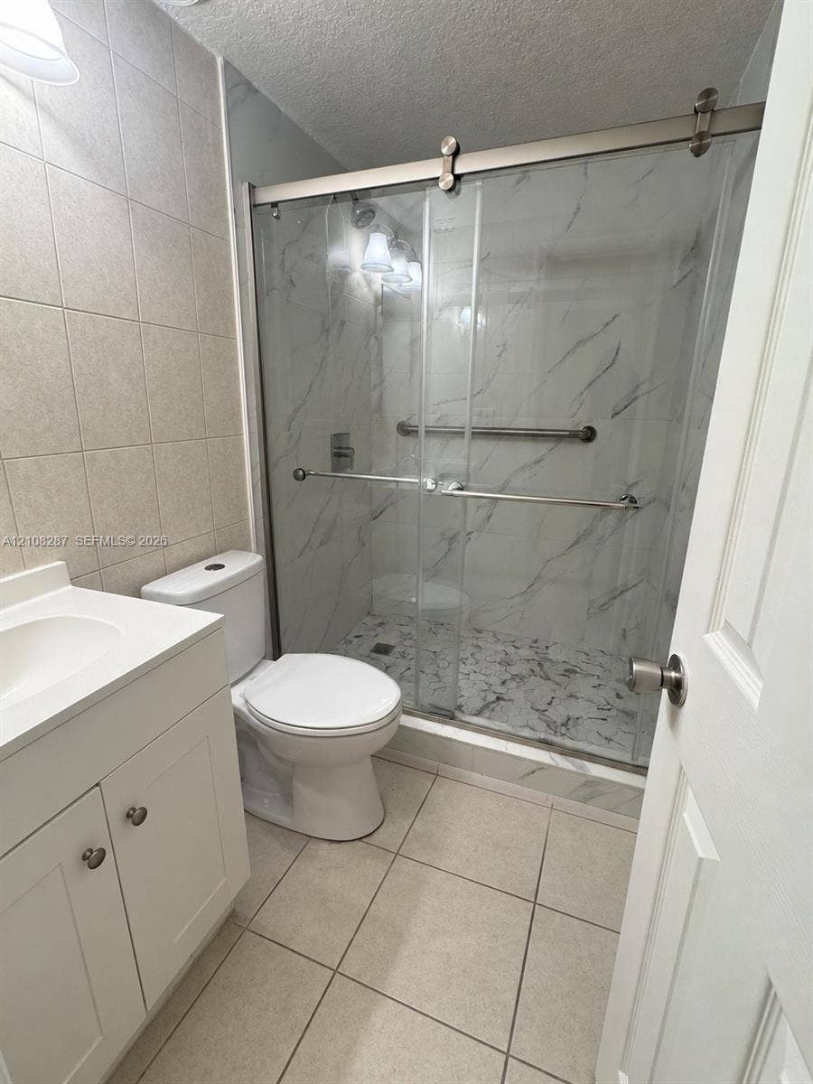 5350 NW 183rd Street, Miami Gardens, FL 33055 Bathroom featuring a white vanity with integrated basin, floor-to-ceiling tile, and a walk-in shower with sliding glass doors, stone-patterned wall surfacing, and metal hardware