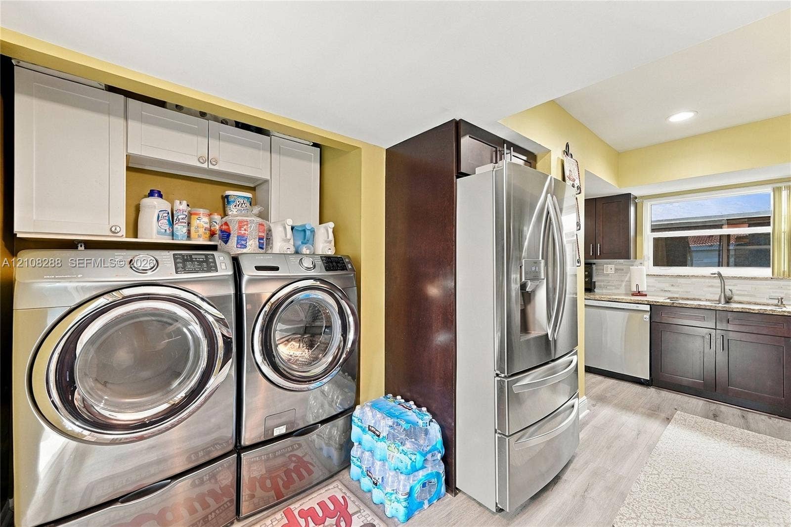 8292 NW 36th Street, Sunrise, FL 33351 Laundry area featuring stainless steel front-load appliances and custom white cabinetry, incorporating a kitchen space with dark-toned cabinetry, stainless steel refrigerator and dishwasher, and stone-surfaced countertops
