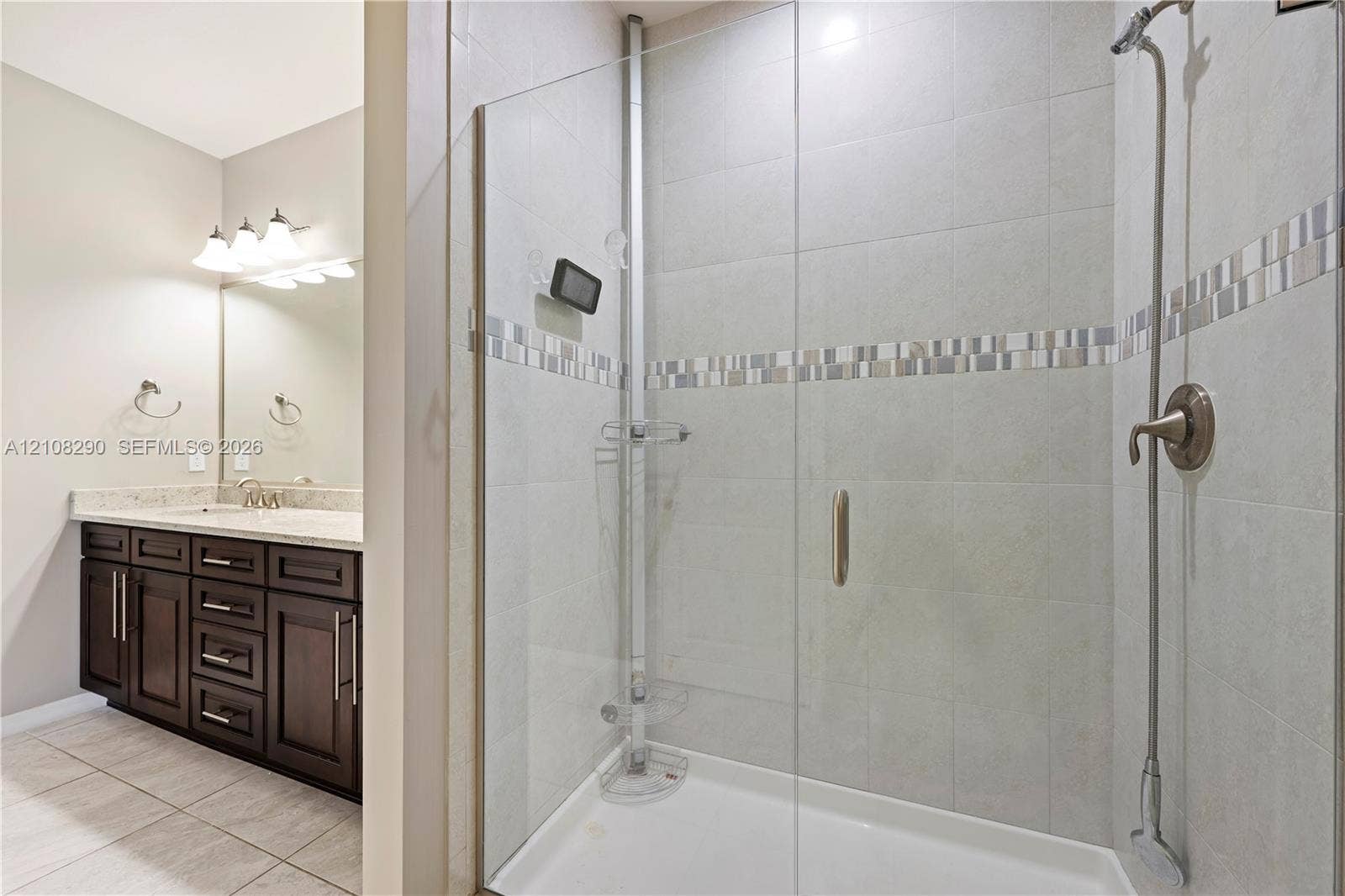 Walk-in shower with a glass enclosure, tile surround, and metallic fixtures, featuring a vanity with stone-finish countertop, dark wood cabinetry, and a full-width wall mirror