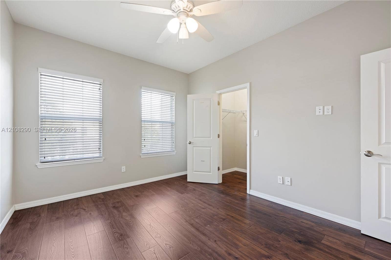 Interior space featuring wood-finish flooring, two windows with horizontal blinds, a ceiling fan with light fixture, and an open doorway to a walk-in closet
