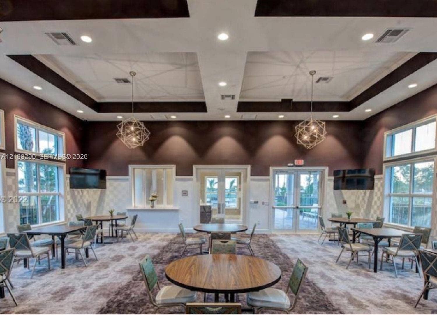 Coffered ceiling with dark accent trim, recessed lighting, dual geometric metal chandeliers, extensive wall-to-wall windows, and double glass-panel entry doors
