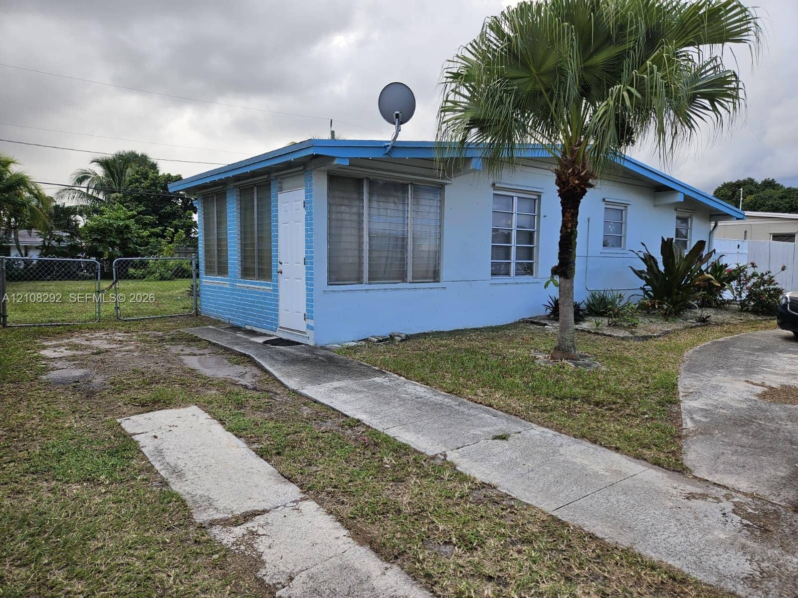 3556 N 10th Avenue N, Palm Springs, FL 33461 Exterior featuring painted brick siding, multi-pane windows, a concrete driveway, a paved walkway, and an enclosed chain-link fenced lot