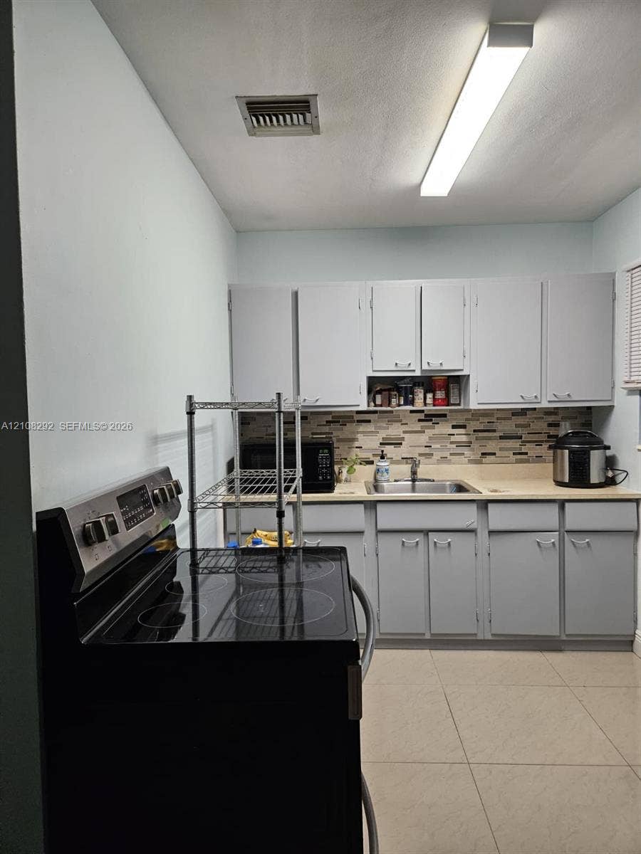 3556 N 10th Avenue N, Palm Springs, FL 33461 Kitchen featuring gray cabinetry, a multi-tone tile backsplash, a stainless steel sink with a mounted faucet, and large-format floor tiling