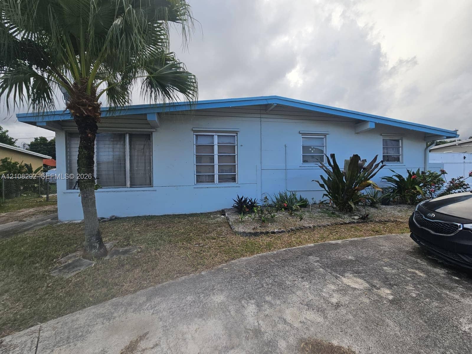 3556 N 10th Avenue N, Palm Springs, FL 33461 Single-story exterior featuring light blue siding, a low-pitch roofline, exposed rafter tails, metal-frame windows, and a concrete driveway