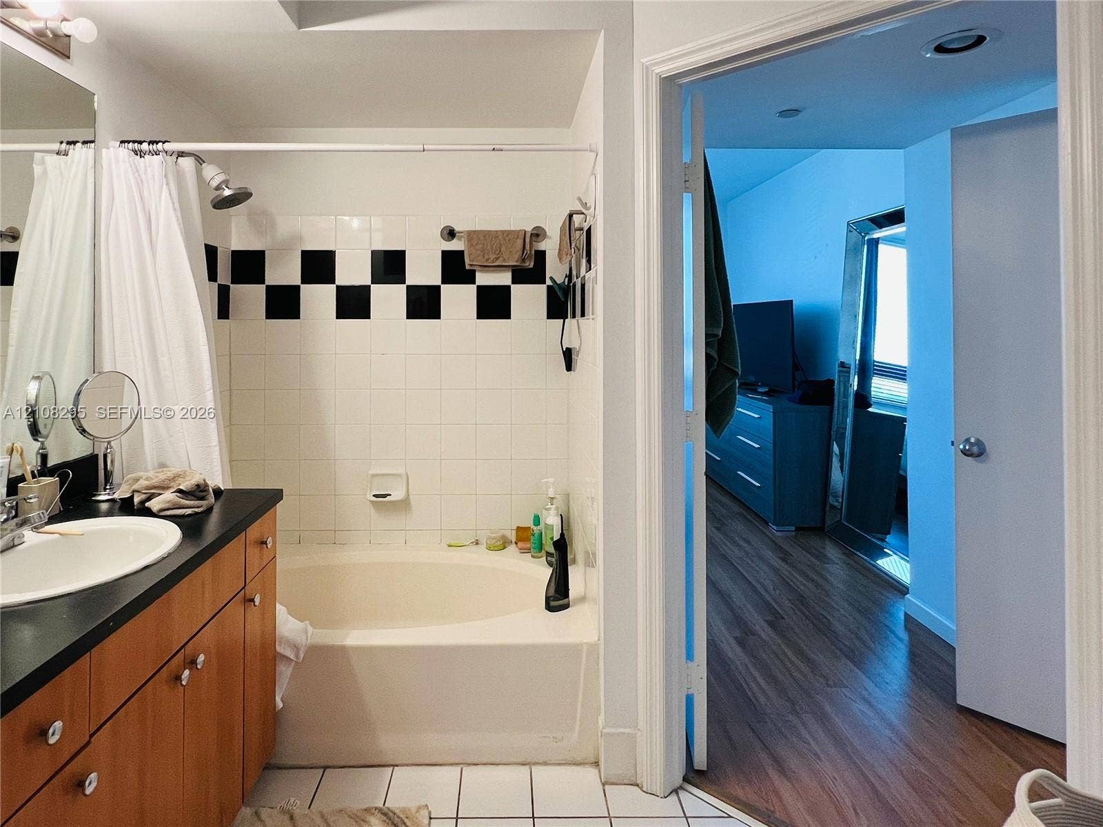 Bathroom featuring a wood-finish vanity with a dark countertop and integrated sink, a soaking tub with a tiled surround featuring a black and white checkerboard border, and white ceramic floor tile