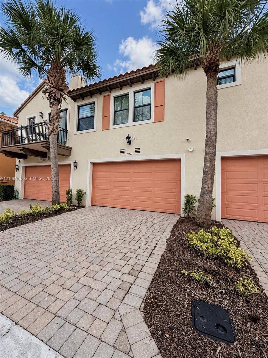 Stucco exterior featuring a dual-bay garage, a second-story balcony with metal railings, tile roof accents, and paver driveway