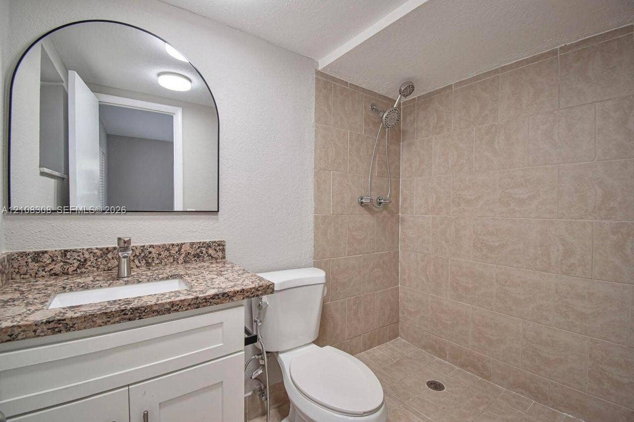 770 SE 2nd Avenue, #E201, Deerfield Beach, FL 33441 Bathroom featuring a white vanity with a stone-finish countertop, undermount sink, arched wall mirror, and walk-in shower with neutral tile surround and adjustable handheld showerhead