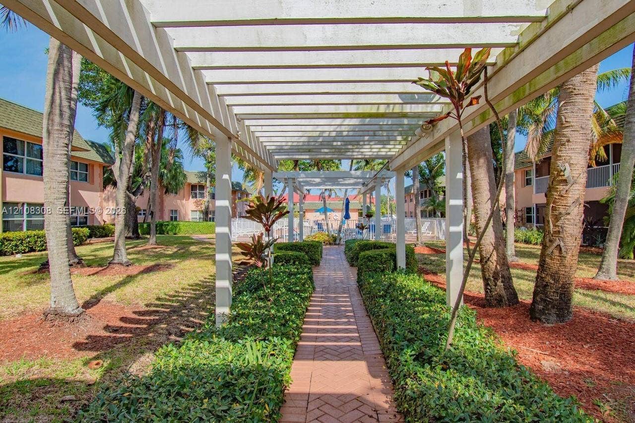 770 SE 2nd Avenue, #E201, Deerfield Beach, FL 33441 White wooden pergola extending over a brick-paved walkway, bordered by manicured hedge rows and mature palm trees
