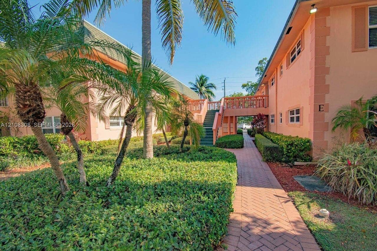 770 SE 2nd Avenue, #E201, Deerfield Beach, FL 33441 Exterior facade featuring peach-toned stucco, brick-style paver walkways, mature palm trees, sculpted hedges, and a raised exterior staircase with balustrades