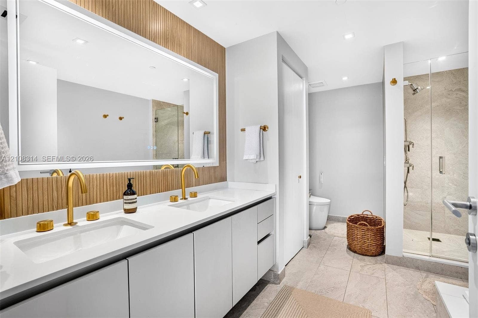 488 NE 18th Street, #4010, Miami, FL 33132 Dual-sink vanity with white surface, metallic gold-finish faucets, large-scale vanity mirror with integrated lighting, wood-slat wall paneling, and walk-in shower with glass enclosure