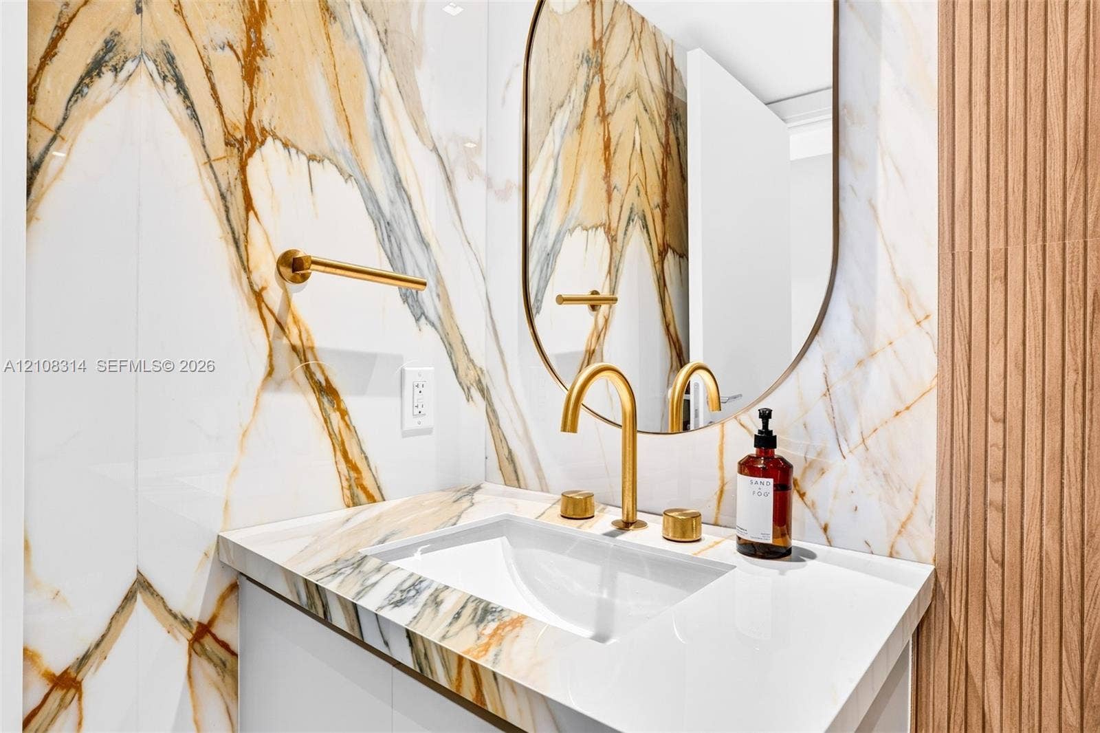 488 NE 18th Street, #4010, Miami, FL 33132 Wall-mounted vanity featuring large-format stone-patterned tile wall cladding, an integrated undermount sink, brass-finish widespread faucet, arched vanity mirror, and wood-slat accent paneling