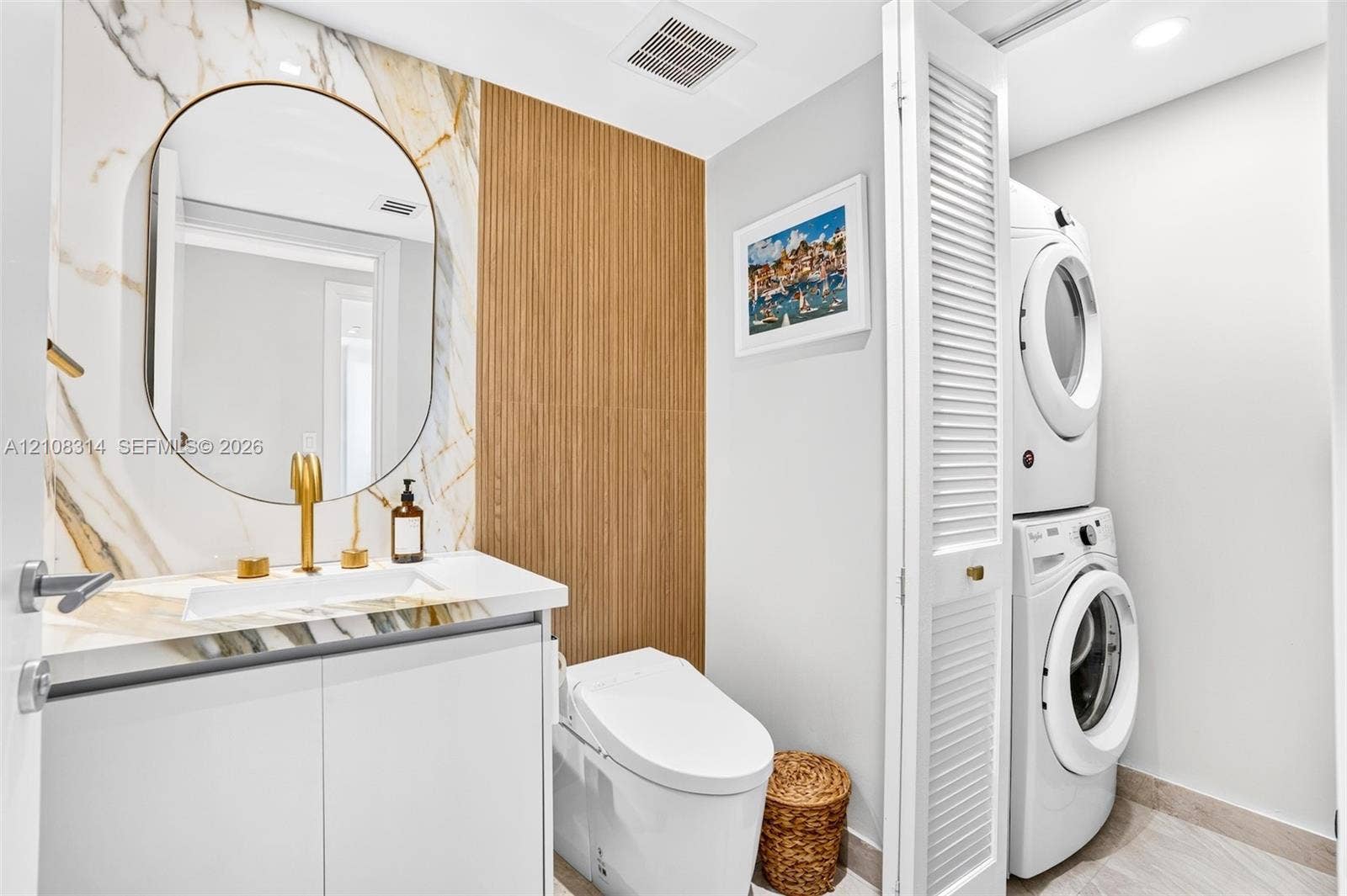 488 NE 18th Street, #4010, Miami, FL 33132 Stone-surround vanity with a circular mirror and gold-tone faucet, complemented by vertical slat wood-finish wall paneling, a low-profile toilet, and a louvered-door closet containing a stacked laundry unit