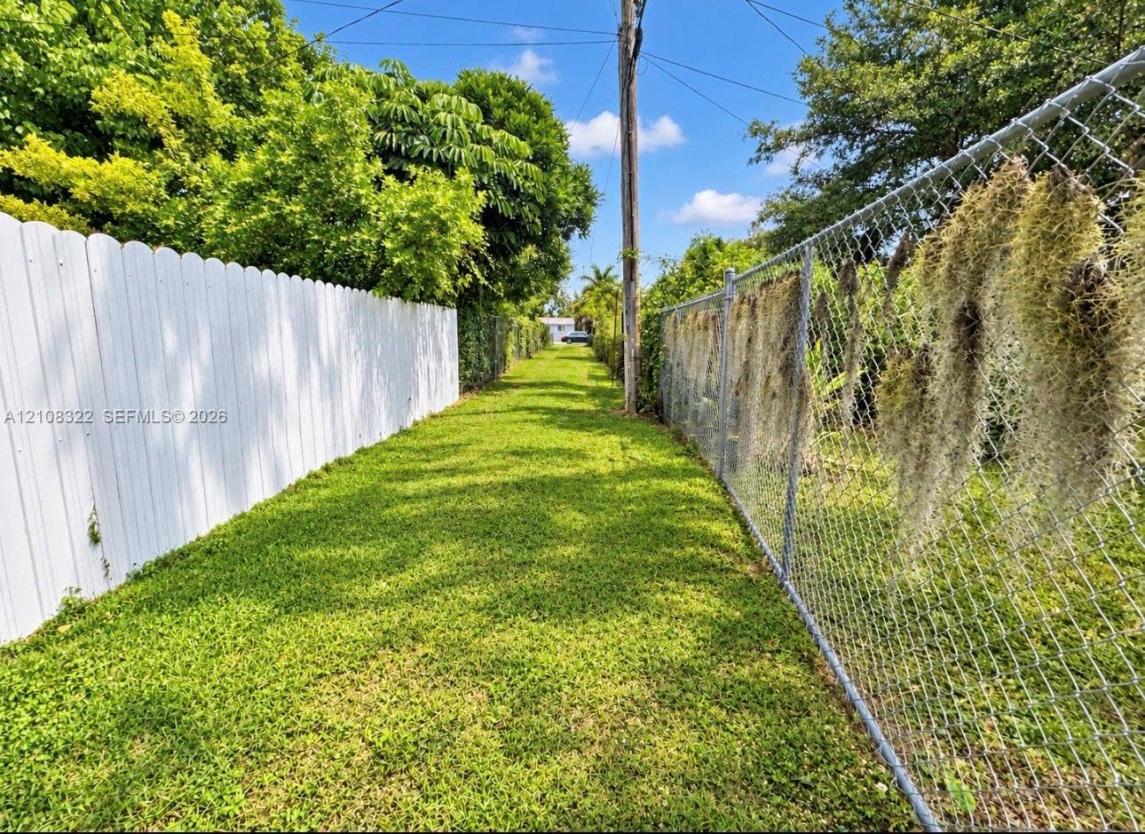 722 NW 7th Avenue, Homestead, FL 33030 Expansive side yard featuring a manicured lawn, a solid white privacy fence, and a chain-link fence perimeter with mature trees
