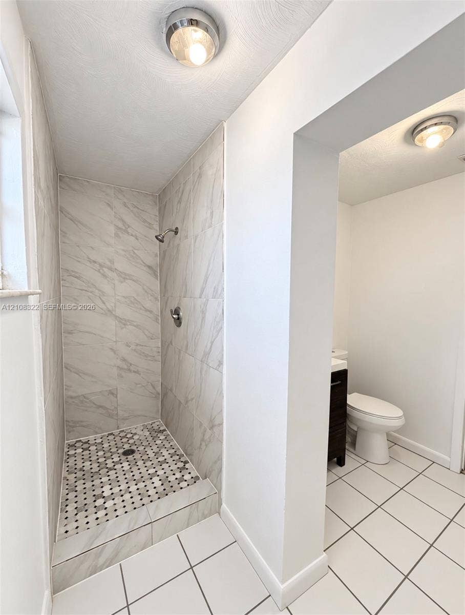 722 NW 7th Avenue, Homestead, FL 33030 Shower stall featuring large-format light-toned stone-finish wall tile, a decorative hexagonal mosaic tile floor, and a polished metal shower fixture, paired with a separate water closet area featuring large-format white tile flooring