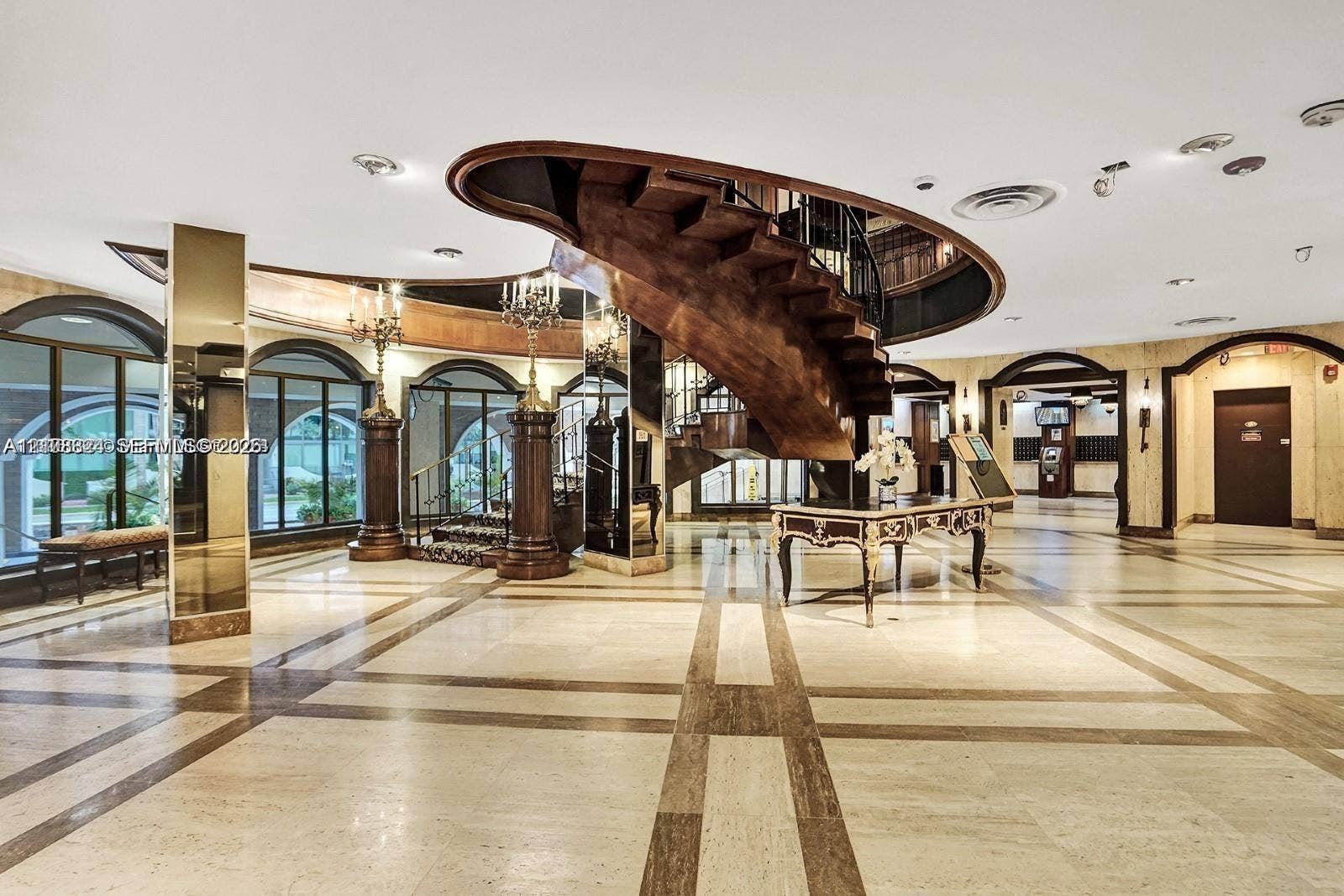 6039 Collins Avenue, #1208, Miami Beach, FL 33140 Grand lobby featuring a suspended spiral wooden staircase, polished stone tile flooring with decorative inlay, arched entryways, mirrored support columns, and recessed ceiling lighting