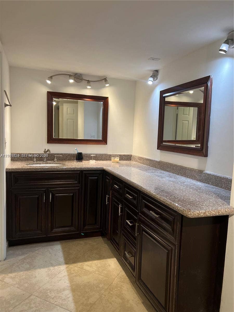 6039 Collins Avenue, #1208, Miami Beach, FL 33140 L-shaped vanity featuring dark wood-finish cabinetry, stone-composite countertops, dual framed mirrors, and multi-bulb track lighting