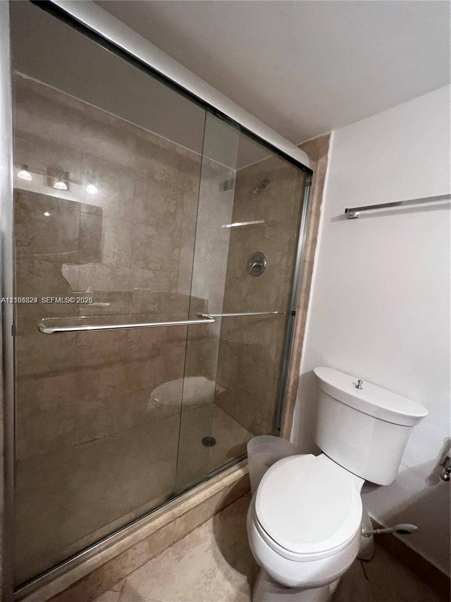 6039 Collins Avenue, #1208, Miami Beach, FL 33140 Walk-in shower with a sliding glass door, neutral-toned stone-finish tile surround, and chrome-finish fixtures, complemented by a white ceramic toilet and a wall-mounted metal towel rack
