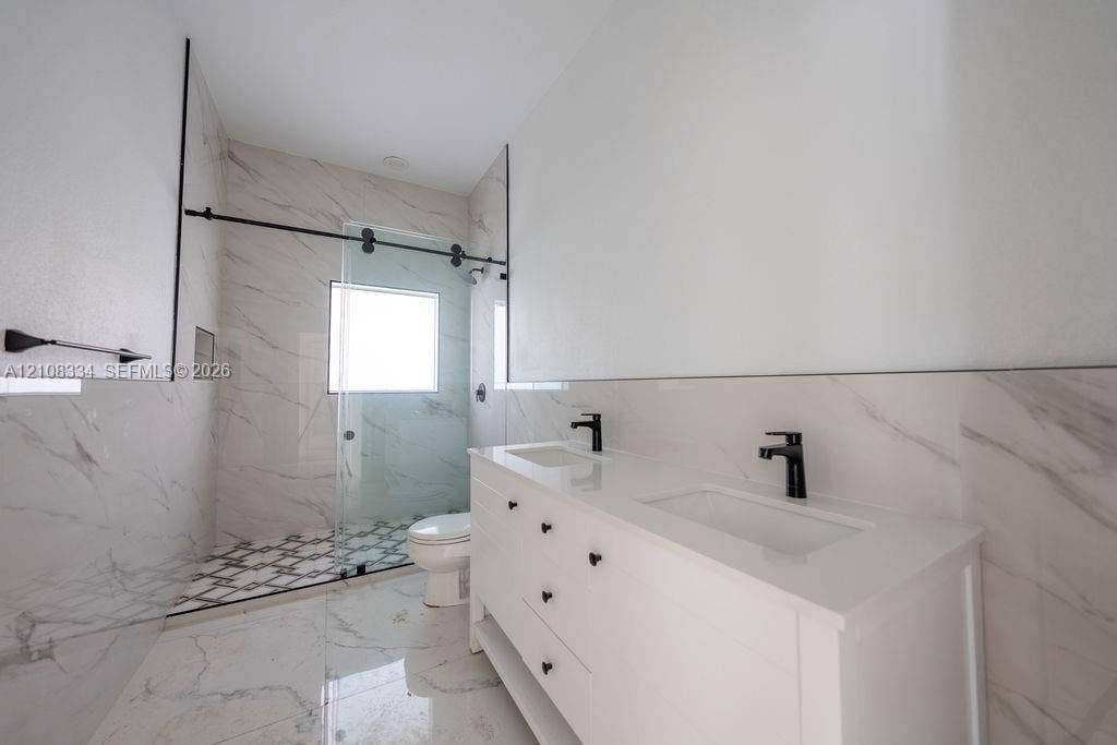Dual-sink vanity with a white countertop and matte black fixtures, walk-in shower with a glass sliding door, large-format stone-patterned tile flooring, and floor-to-ceiling tiled shower surround