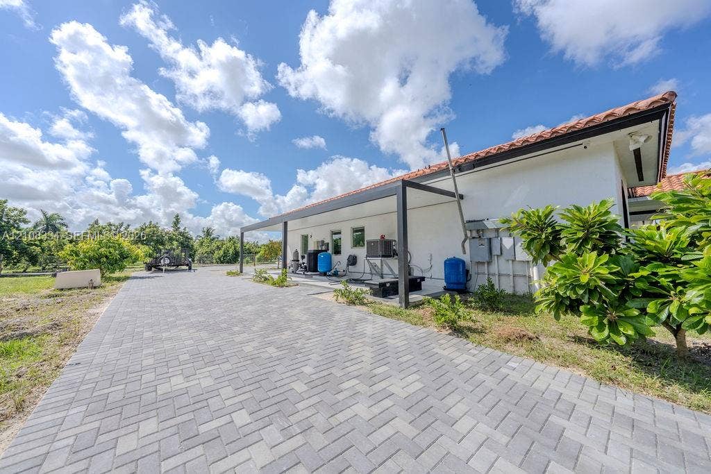 Stucco exterior with a terracotta-style tile roof, expansive gray paver driveway, and a black metal-frame covered side patio