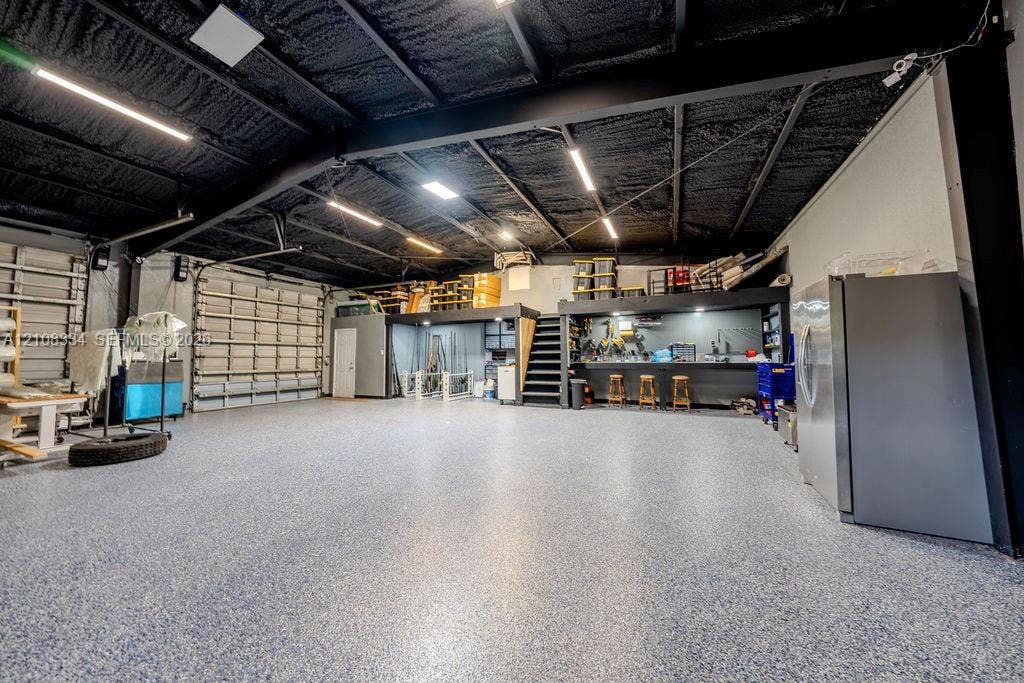 Expansive workshop featuring an epoxy-coated floor, vaulted ceiling with exposed steel beams, built-in workbench, integrated overhead linear lighting, and a mezzanine storage level
