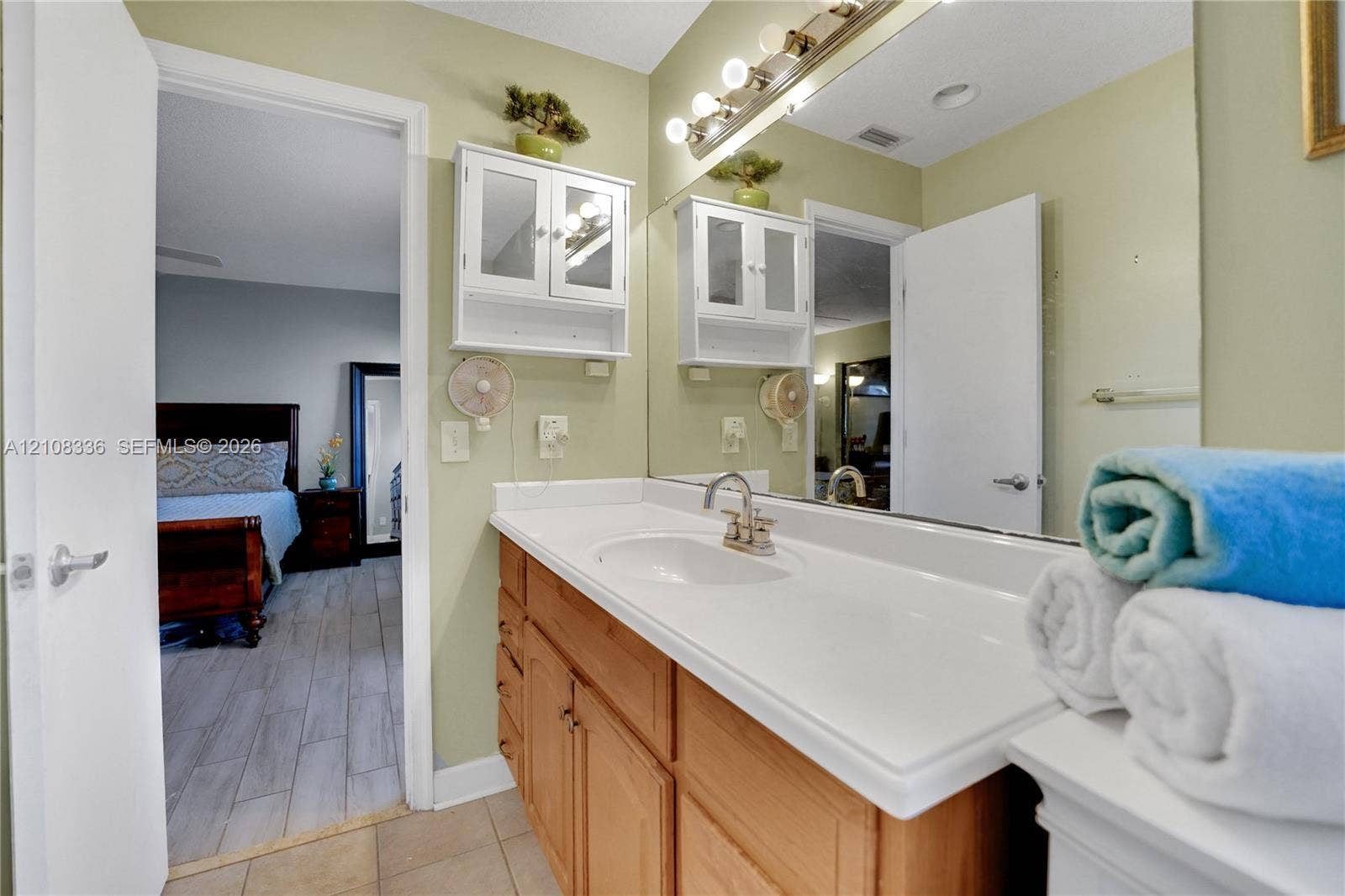 6311 Hitchin Post Way, Delray Beach, FL 33484 Single-sink vanity with a solid-surface countertop, natural wood-finish cabinetry, wall-mounted white storage hutch, and large-scale vanity mirror with integrated multi-bulb lighting