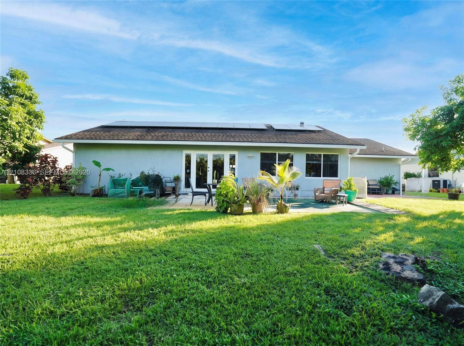 6311 Hitchin Post Way, Delray Beach, FL 33484 Exterior rear elevation featuring a stucco facade, rooftop solar panel array, multi-panel glass door, and a paved patio area set within a spacious lawn