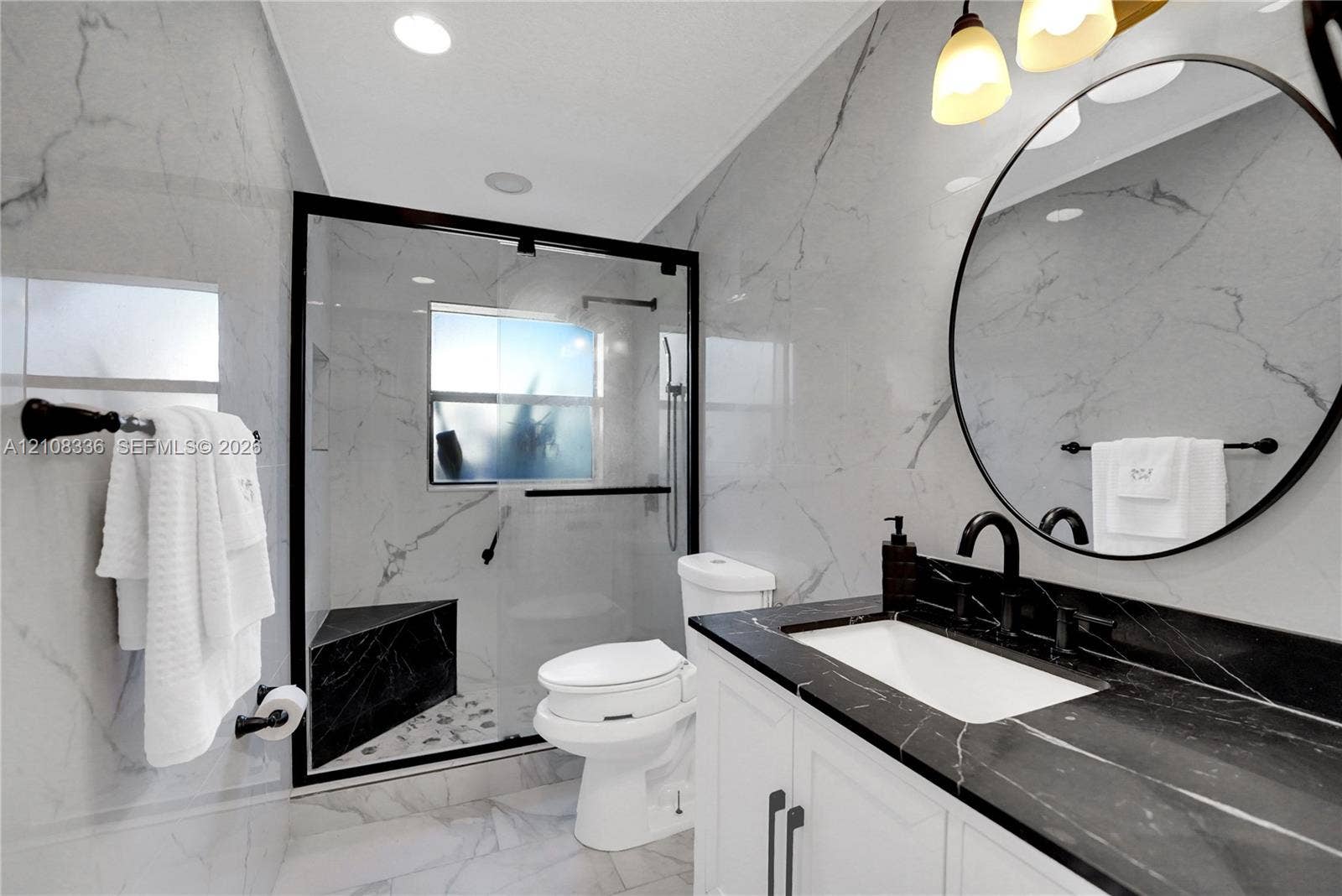 6311 Hitchin Post Way, Delray Beach, FL 33484 Bathroom featuring large-format wall tiling, a walk-in glass-enclosed shower with a built-in bench, a custom vanity with a contrasting dark stone countertop, a circular wall-mounted mirror, and matte black hardware