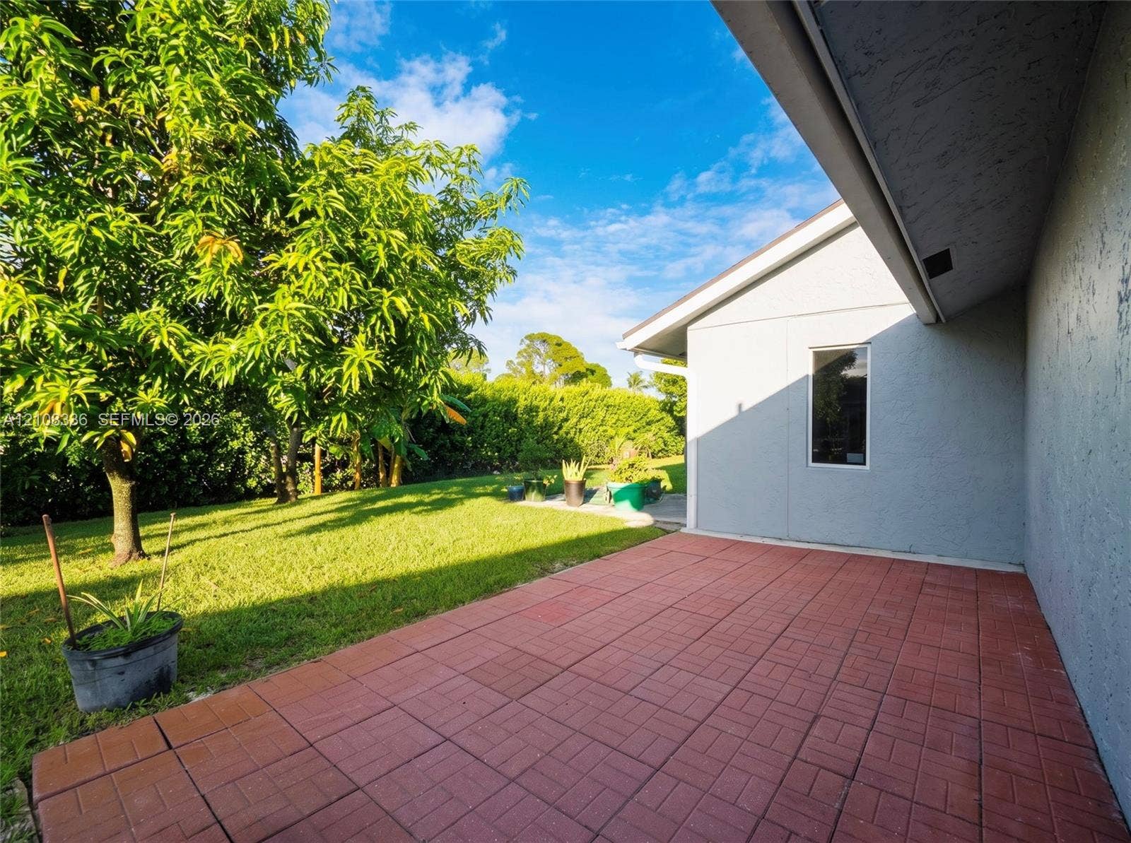 6311 Hitchin Post Way, Delray Beach, FL 33484 Rear exterior featuring a red brick-patterned paved patio, stucco siding, and a landscaped backyard with mature trees