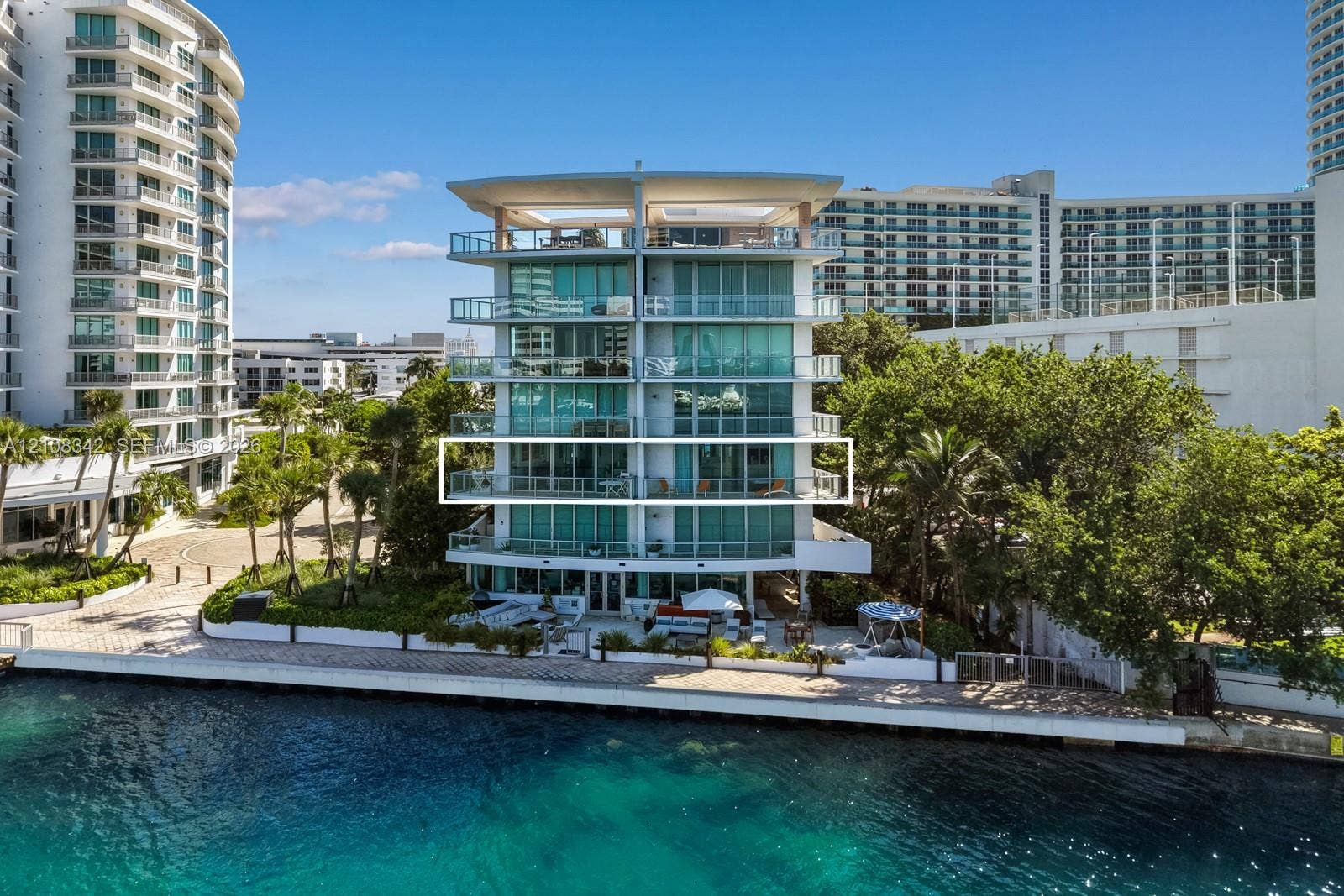 Mid-rise condominium exterior featuring floor-to-ceiling glass windows, wraparound balconies with glass railings, and waterfront hardscaping