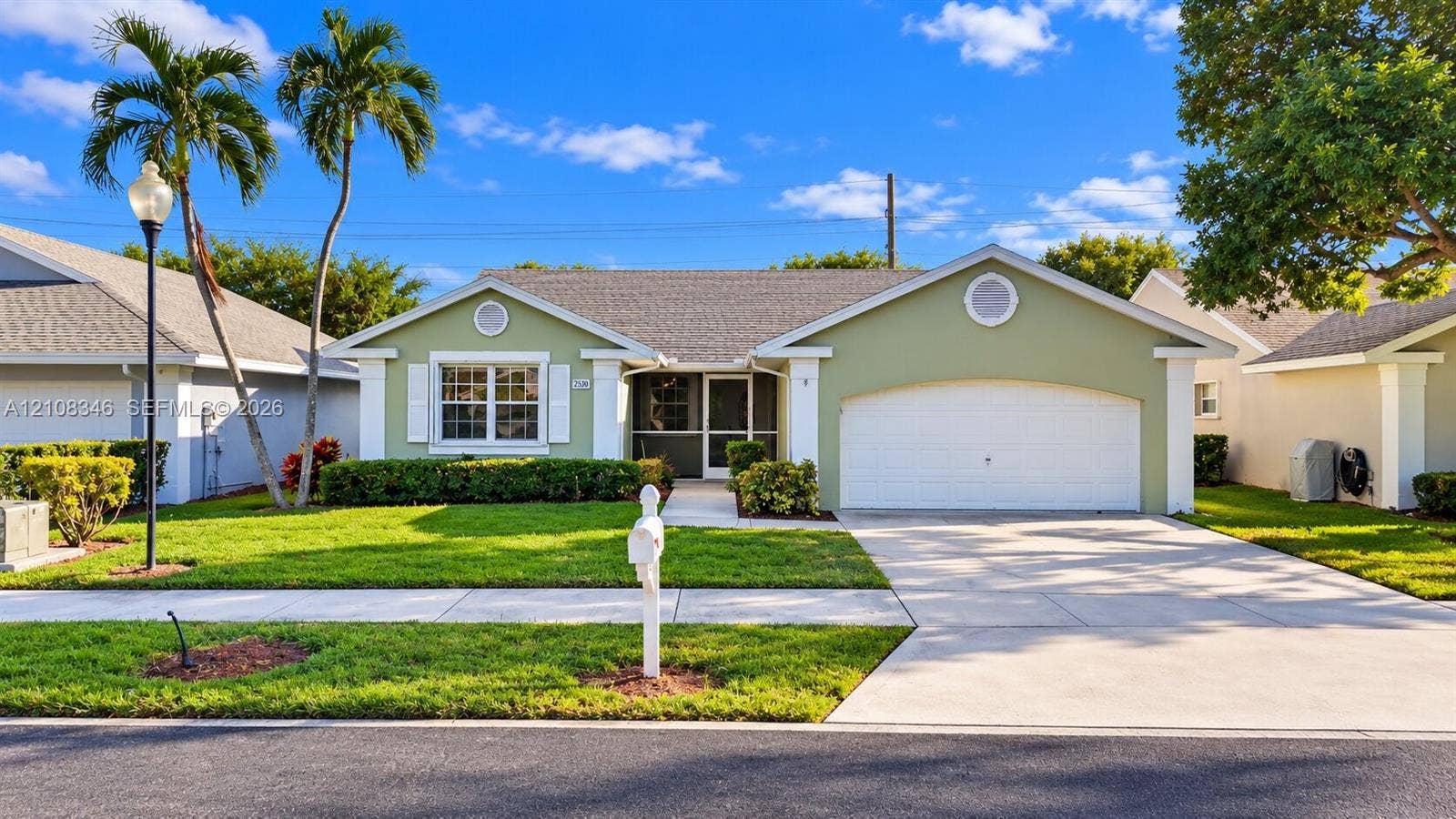 2530 SE 7th Place, Homestead, FL 33033 Single-story exterior featuring a gabled roofline, light green siding with white trim, a multi-car garage, and a manicured lawn with mature palm trees