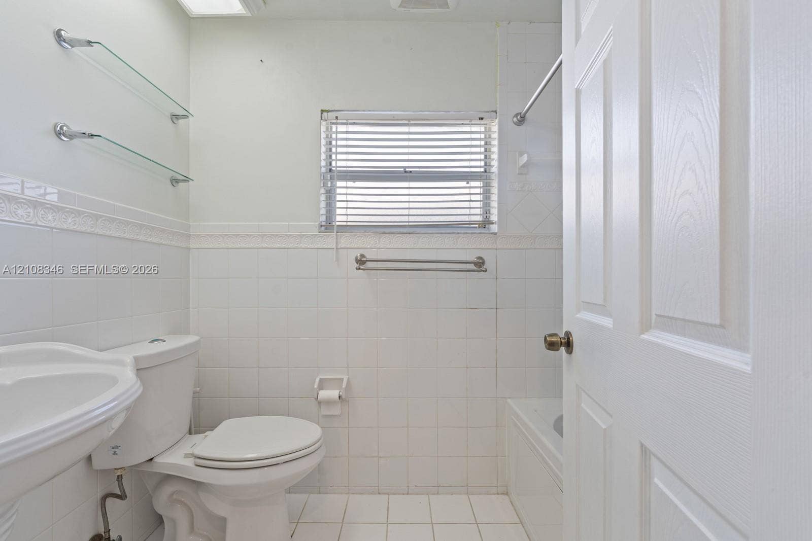 2530 SE 7th Place, Homestead, FL 33033 Bathroom featuring white ceramic wall and floor tiling, a pedestal sink, a built-in bathtub with a metal shower rod, and two integrated glass shelving units