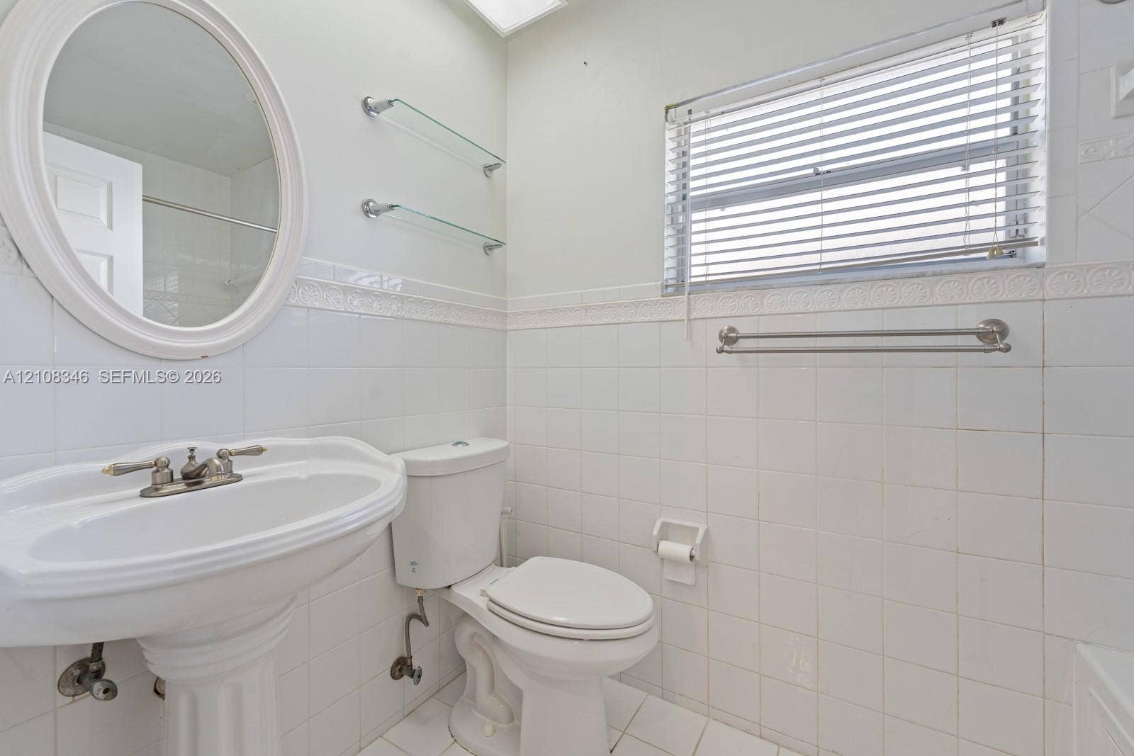 2530 SE 7th Place, Homestead, FL 33033 Bathroom featuring white ceramic tile wall cladding, pedestal sink with polished metal faucet, oval wall-mounted mirror, glass shelving, and horizontal window blinds