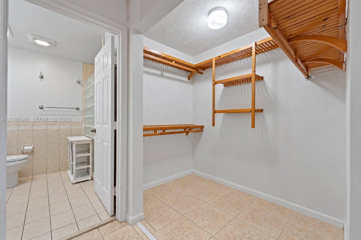 2530 SE 7th Place, Homestead, FL 33033 Walk-in closet featuring multi-level wooden shelving and tile flooring, with an entry to a tiled bathroom equipped with a glass block wall section and mounted hardware
