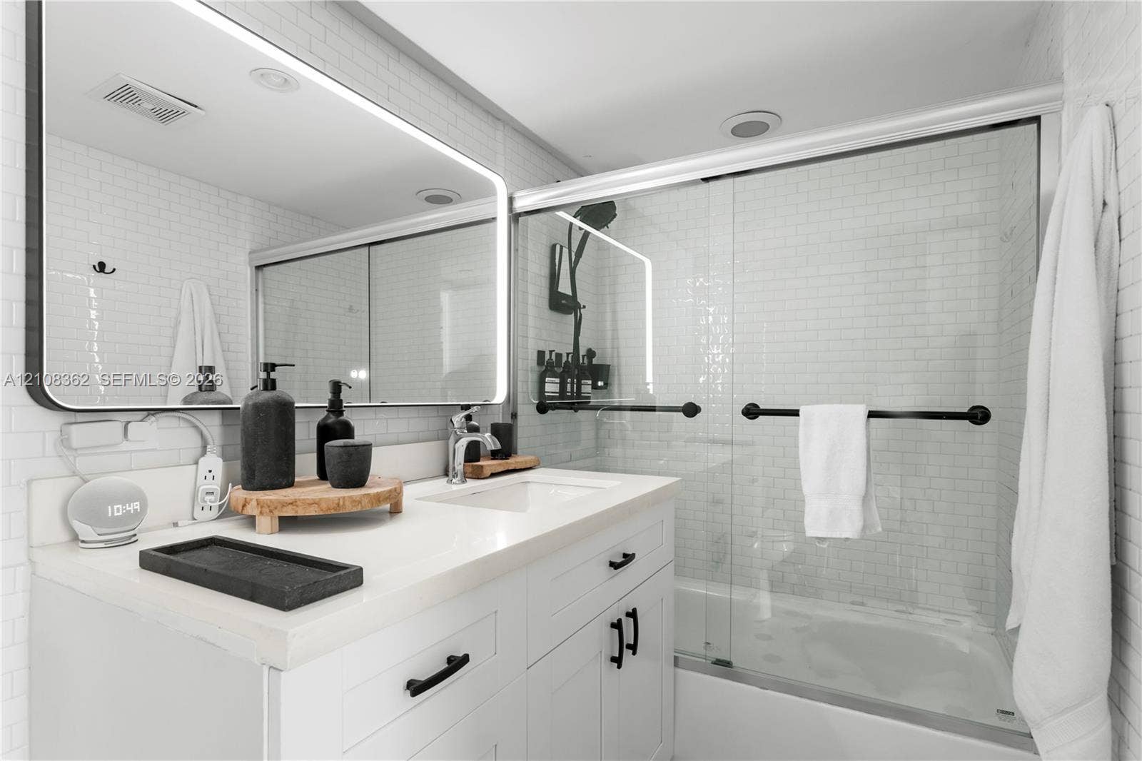 White vanity featuring a solid surface countertop, undermount sink, and matte black hardware, paired with a subway tile shower surround, sliding glass door, and a backlit rectangular mirror