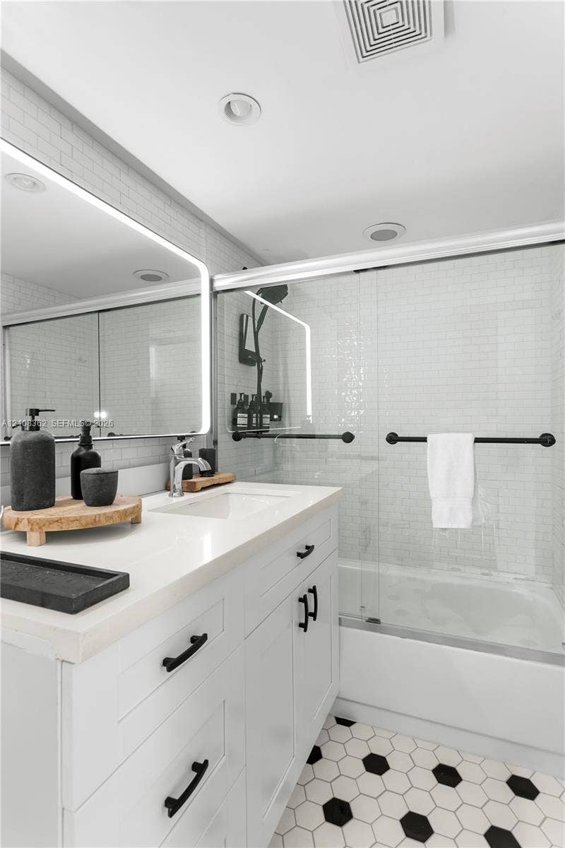 White shaker-style vanity with a solid surface countertop, undermount sink, integrated LED-backlit mirror, bathtub with glass sliding doors, and hexagonal floor tile