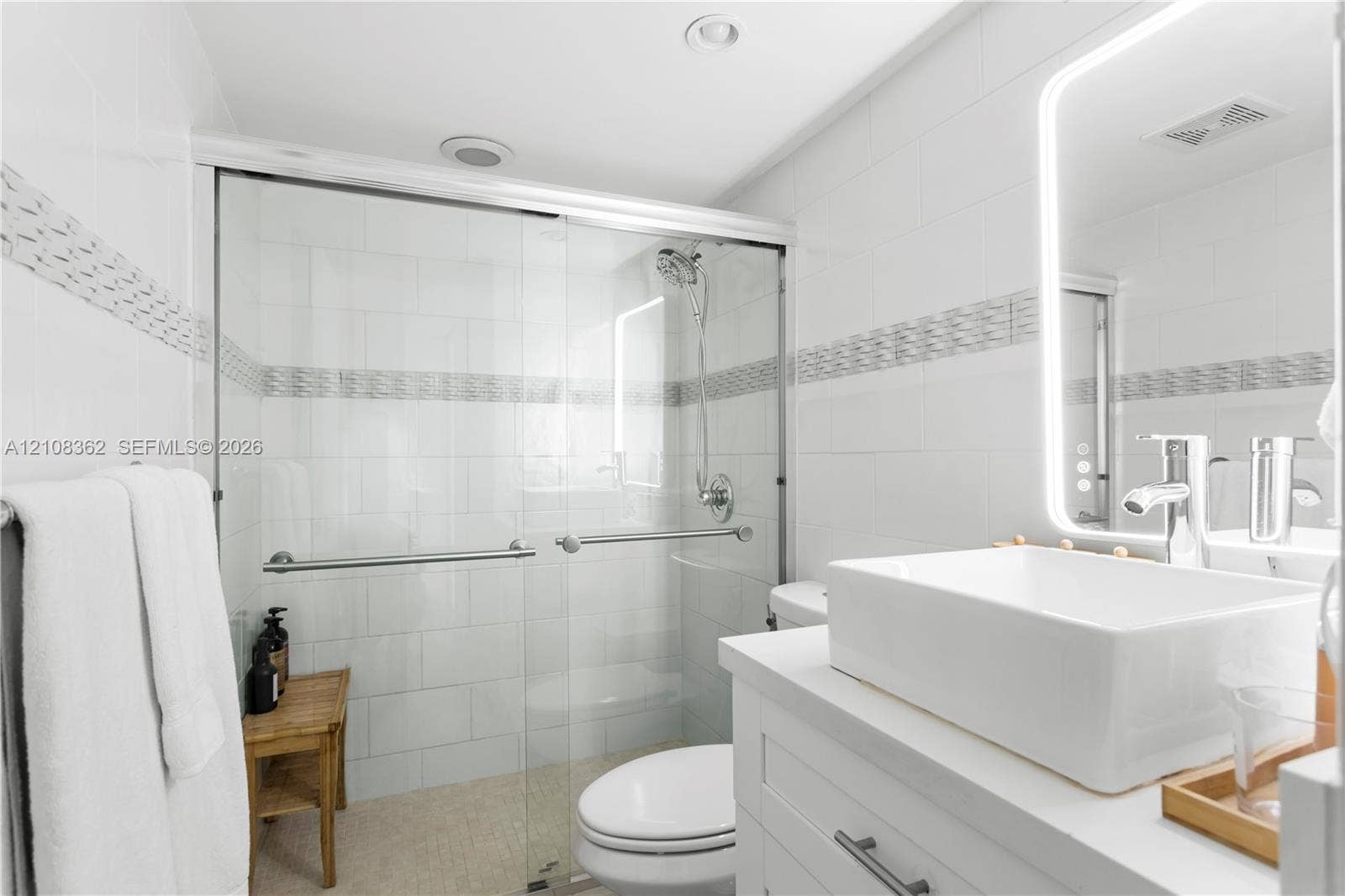 Walk-in shower with sliding glass doors, white subway-style wall tiling, decorative mosaic tile inlay, modern vessel sink with chrome faucet, and integrated LED backlit mirror