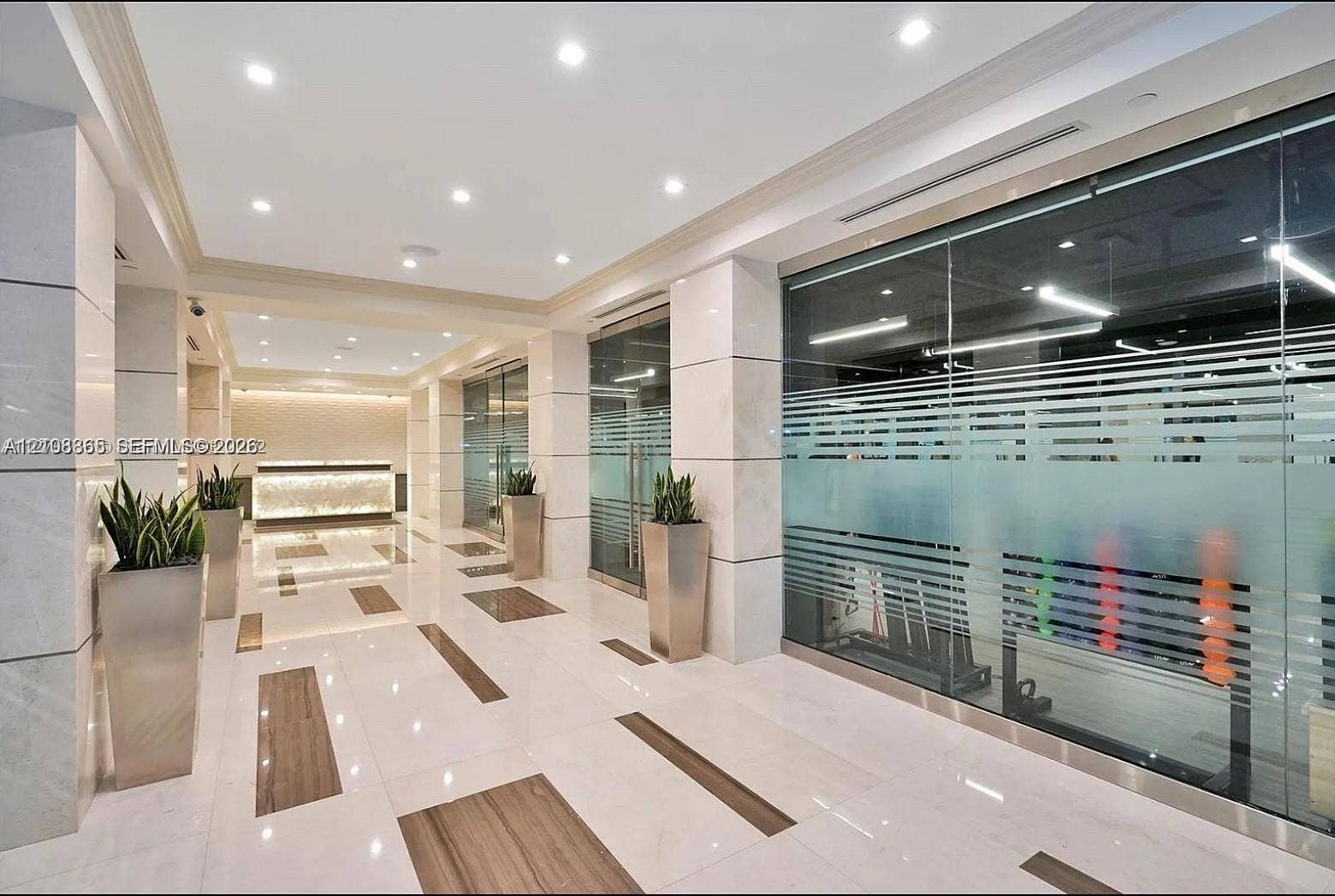 20165 NE 39 Place, #403, Aventura, FL 33180 Polished stone flooring with inlaid wood-finish accents, large-format stone pillar cladding, glass-partitioned wall panels, recessed lighting, and a backlit stone-front desk
