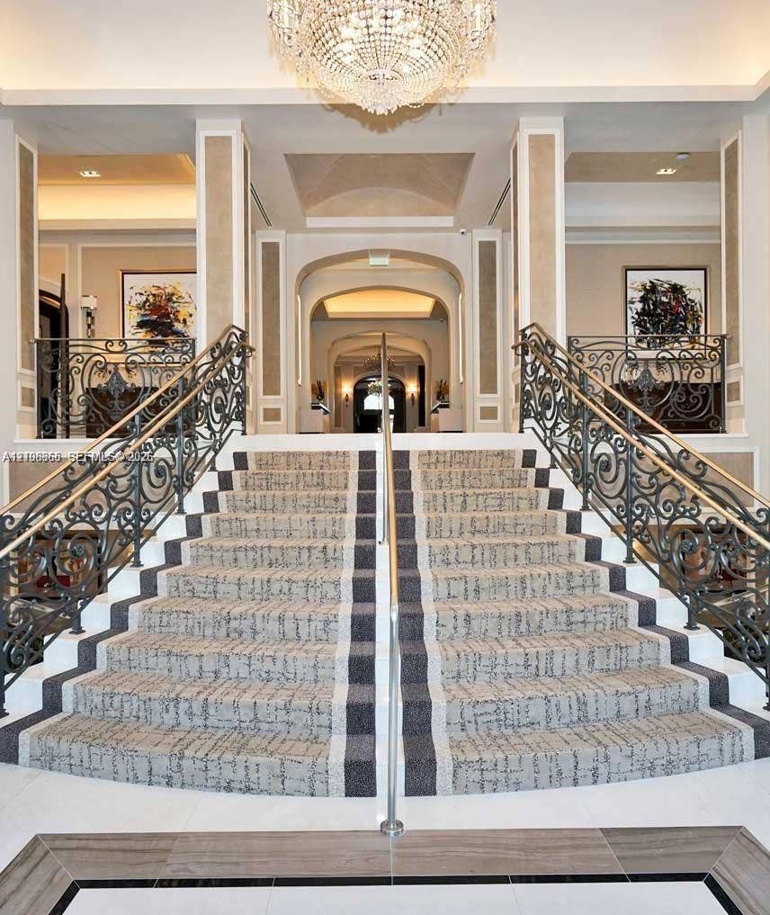 20165 NE 39 Place, #403, Aventura, FL 33180 Grand dual staircase featuring intricate wrought iron railings, stone flooring with decorative inlay, and a central crystal chandelier under vaulted ceiling detailing